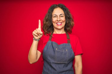 Kırmızı izole edilmiş arka planda önlük giyen orta yaşlı bir kadın bir numaralı parmağı gösterirken kendinden emin ve mutlu bir şekilde gülümsüyor..