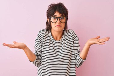 İzole edilmiş pembe arka plan üzerinde çizgili tişört ve gözlük takan genç ve güzel bir kadın. Kolları ve elleri havada, kafası karışmış bir ifade. Şüphe kavramı.