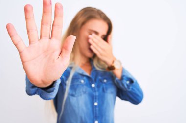 İzole edilmiş beyaz arka planda dikilen sıradan bir kot gömlek giyen genç ve güzel bir kadın ellerle gözlerini kapatıyor ve üzgün ve korku ifadeleriyle dur hareketi yapıyor. Utanç verici ve olumsuz bir kavram..