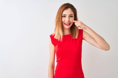 İzole edilmiş beyaz arka planda duran zarif kırmızı elbiseli kızıl saçlı iş kadını. El parmağı ve burunla işaret ediyor, neşeli bir şekilde gülümsüyor. Güzellik konsepti