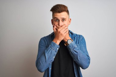 İzole edilmiş beyaz arka plan üzerinde duran kot gömlek giyen genç yakışıklı adam yanlışlıkla elleriyle ağzını kapatırken şok oldu. Gizli konsept.