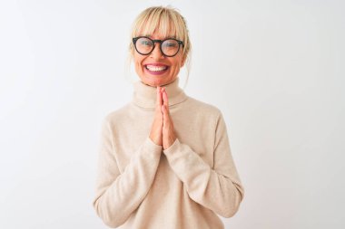 Balıkçı yaka kazak giyen ve gözlüklü orta yaşlı bir kadın izole edilmiş beyaz arka plan üzerine dua ederek ellerini birleştirip af diliyor kendinden emin bir şekilde gülümsüyor.