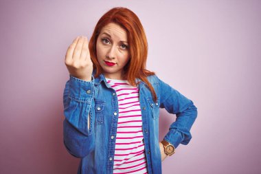Güzel kızıl saçlı kadın kot gömlek ve çizgili tişört giyerek izole edilmiş pembe arka planda İtalyan jesti yapıyor.