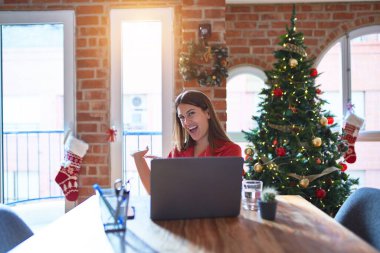 Güzel bir kadın evde dizüstü bilgisayarla çalışıyor Noel ağacının etrafında elini açıp gülümseyerek içeri girmeye davet ediyor.