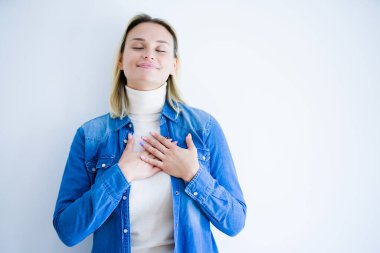 İzole edilmiş beyaz arka planda duran kot gömlek giyen genç ve güzel bir kadın. Elleri göğsünde, gözleri kapalı ve yüzünde minnet dolu bir jest ile gülümsüyor. Sağlık kavramı.