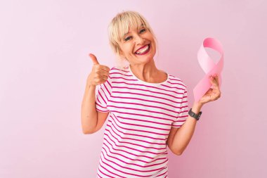Kanser kurdelesi tutan orta yaşlı bir kadın pembe arka planda duruyor. Kocaman gülümsüyor ve gülümsüyor. İşaret veriyor.