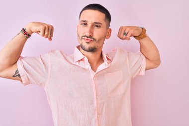 Arkaplanda duran pembe tişörtlü yakışıklı genç adam gururla gülümseyen kol kaslarını gösteriyor. Fitness konsepti.