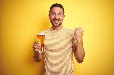 Genç yakışıklı adam izole edilmiş sarı arka planda bira içerken gururla bağırıyor ve zaferi ve başarıyı kutluyor. Çok heyecanlı, coşkulu bir duygu.