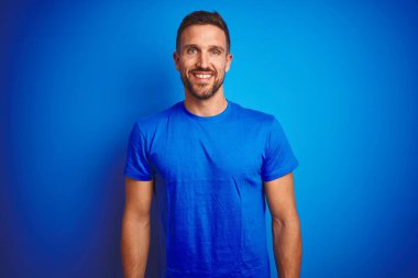 Mavi izole edilmiş arka plan üzerine sıradan tişört giyen genç yakışıklı bir adam. Yüzünde mutlu ve havalı bir gülümseme var. Şanslı kişi..