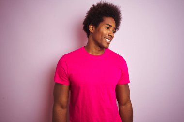 Afro saçlı genç Amerikalı adam izole edilmiş pembe arka planda dikilip yüzünde gülümseme, doğal bir ifade ile bir o yana bir bu yana bakıyor. Kendinden emin gülen.