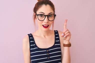 Kırmızı saçlı, at kuyruklu, çizgili tişörtlü, pembe arka planda duran bir kadın bir fikir ya da soru işareti ile şaşırmış, mutlu yüzlü, bir numara