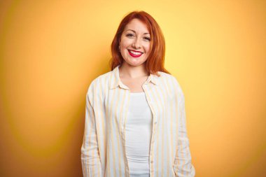 Güzel, kızıl saçlı, çizgili gömlek giyen, izole edilmiş sarı arka plan üzerinde duran mutlu ve havalı bir gülümsemesi olan bir kadın. Şanslı kişi..