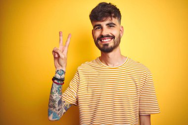 İzole edilmiş sarı arka planda duran çizgili tişörtlü genç adam kameraya bakıp zafer işareti yaparken gülümsüyor. İki numara..