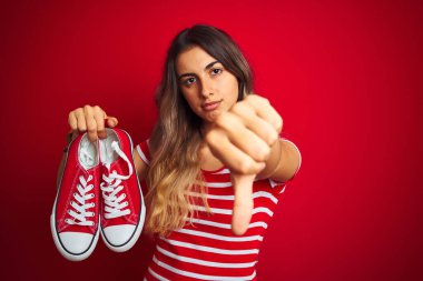 Güzel bir kadın, kırmızı izole edilmiş arka planda spor ayakkabıları tutuyor. Kızgın bir yüzü var. Negatif işaretler, başparmakları aşağıda, reddedilme kavramından hoşlanmadığını gösteriyor.