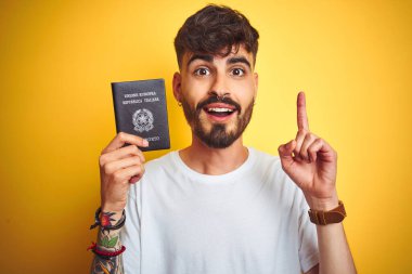 İtalyan pasaportu takan dövmeli genç bir adam izole edilmiş sarı arka planda bir fikir ya da soru işareti ile şaşırdı.