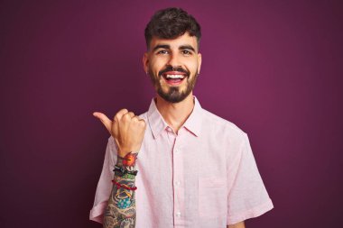 Yalıtılmış mor arka planda duran pembe tişörtlü, dövmeli genç adam gülümseyen yüz ifadesiyle başparmağıyla yan tarafa bakıyor ve işaret ediyor..