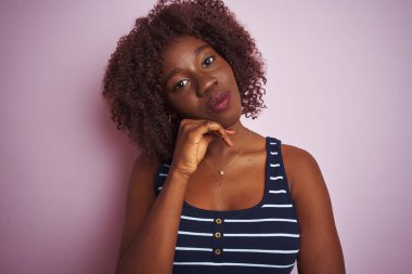 Çizgili tişört giyen Afrikalı genç bir kadın pembe arka planda duruyor ve elleri çenesinde soru sormayı düşünüyor. Düşünceli bir ifade. Düşünceli bir yüzle gülümsüyor. Şüphe kavramı.