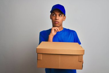 Genç yakışıklı arap teslimat adam izole beyaz arka plan üzerinde duran bir kutu tutan ciddi yüz soru hakkında düşünme, çok karışık bir fikir