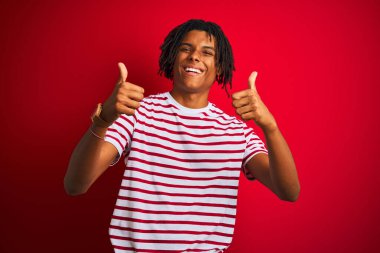 Rastalı tişörtlü genç afro adam izole edilmiş kırmızı arka plan işareti üzerinde duruyor. Elleriyle olumlu jest yapıyor. Gülümsüyor ve mutlu. Neşeli ifade ve kazanan jest.