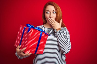 Kızıl saçlı güzel bir kadın, kırmızı izole edilmiş arka plan üzerinde doğum günü hediyesini tutuyor. Ağzı kapalı, utancından ağzı açık, korku ifadesi, sessizlikten korkmak, gizli bir konsept.