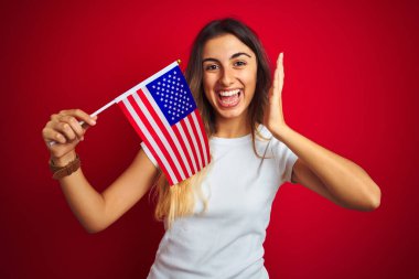 Amerika Birleşik Devletleri bayrağını kırmızı izole arka planda tutan genç bir kadın çok mutlu ve heyecanlı, kazanan ifade zaferi büyük bir gülümsemeyle kutluyor ve ellerini kaldırıyor.