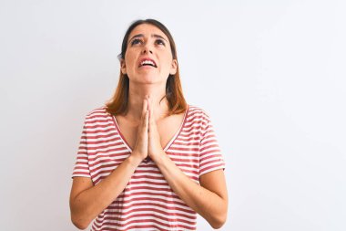 Güzel kızıl saçlı kadın, izole edilmiş arka planda sıradan kırmızı tişörtler giyip elleri bağlı yalvarıyor ve dua ediyor. Yüzünde umut ifadesi var. Çok duygusal ve endişeli. Af diliyor. Din kavramı.