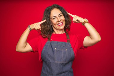 Kırmızı izole edilmiş arka planda önlük giymiş orta yaşlı bir kadın iki eliyle başını gösteren bir gülümseme, harika bir fikir ya da düşünce, iyi bir hafıza