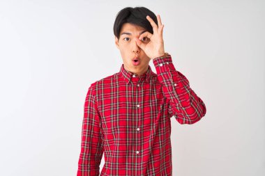 İzole edilmiş beyaz arka plan üzerinde duran sıradan kırmızı tişörtlü genç Çinli adam şaşırmış yüz ifadesiyle şaşırmış, parmaklarının arasından bakan bir hareket yapıyor. İnançsız ifade.