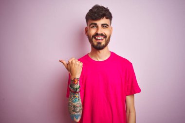 Dövmeli genç adam pembe arka planda dikilip gülümseyen ve baş parmağıyla yüzünü gösteren pembe tişörtlü..