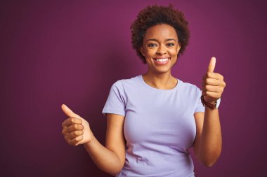 Afro saçlı, güzel, Afro saçlı genç bir Amerikalı kadın. Mor arka planda başarı işareti var. Elleriyle olumlu jest yapıyor. Neşeli ifade ve kazanan jest.