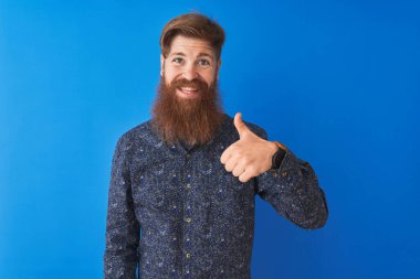 Çiçekli yaz tişörtü giyen genç kızıl saçlı adam izole edilmiş mavi arka planda durup el hareketleriyle mutlu mesut işaret veriyor. Kameraya bakıp başarıyı gösteren ifade onaylanıyor.