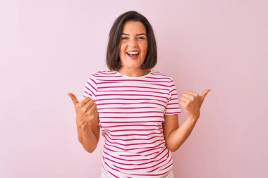 İzole edilmiş pembe arka plan başarısı tabelasının üzerinde duran çizgili tişört giyen genç ve güzel bir kadın. Elleriyle olumlu jest yapıyor. Gülümsüyor ve mutlu. Neşeli ifade ve kazanan jest.