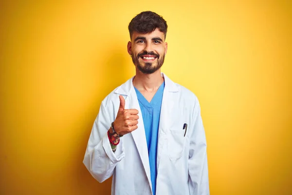 Young doctor man with tattoo standing over isolated yellow background doing happy thumbs up gesture with hand. Approving expression looking at the camera with showing success.