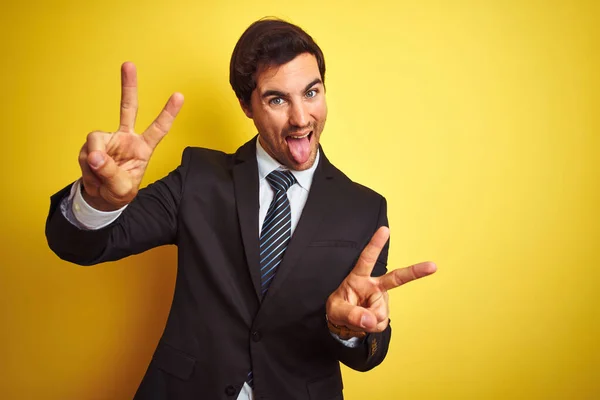 若いハンサムなビジネスマンがスーツを着て 両方の手の勝利の兆候を行うの指を示す舌で笑って孤立した黄色の背景の上に立ってネクタイ 第二番 — ストック写真