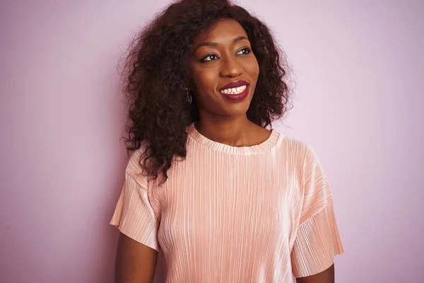 Young african american woman wearing t-shirt standing over isolated pink background looking away to side with smile on face, natural expression. Laughing confident.