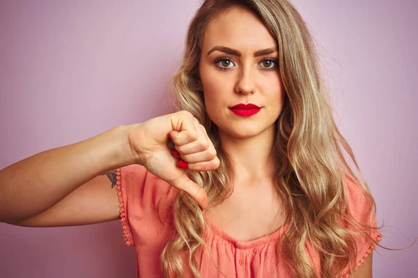 年轻漂亮的女人穿着T恤 站在粉色孤立的背景上 满脸怒容 负面的迹象表明她不喜欢大拇指朝下 拒绝的观念 — 图库照片