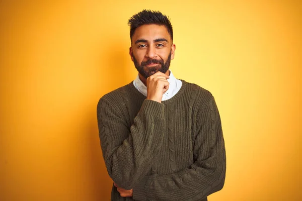 Young indian man wearing green sweater and shirt standing over isolated yellow background looking confident at the camera with smile with crossed arms and hand raised on chin. Thinking positive.