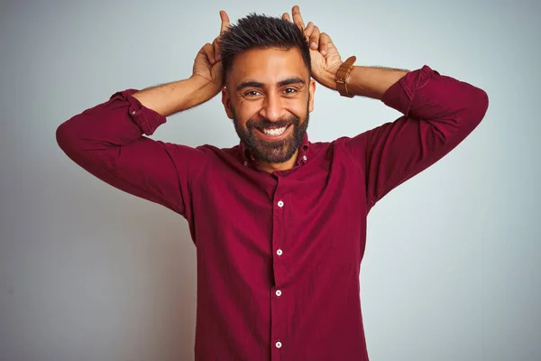 Young indian man wearing red elegant shirt standing over isolated grey background Posing funny and crazy with fingers on head as bunny ears, smiling cheerful