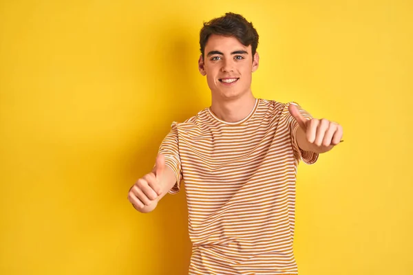 Teenager boy wearing yellow t-shirt over isolated background approving doing positive gesture with hand, thumbs up smiling and happy for success. Winner gesture.