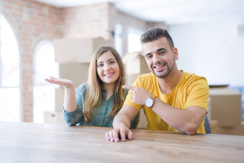Young couple sitting on the table movinto to new home with carboard boxes behind them amazed and smiling to the camera while presenting with hand and pointing with finger.