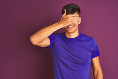 İzole edilmiş mor arka planda duran tişört giyen genç Hintli adam, gülümsüyor ve elleri yüzünde şaşkınlıktan gözleri kapayarak gülüyor. Kör kavram.