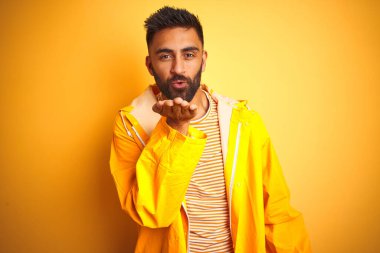 Yağmurluk giyen genç Hintli adam izole edilmiş sarı arka planda duruyor ve kameraya bakıp havada el ele öpüşerek güzel ve seksi bir şekilde öpüşüyorlar. Aşk ifadesi.