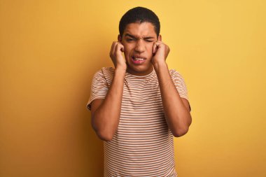 İzole edilmiş sarı arka planda duran çizgili tişört giyen genç, yakışıklı Arap adam yüksek sesli müzik sesi için rahatsız edici ifadeleri olan parmaklarıyla kulakları kapatıyor. Sağır kavramı.