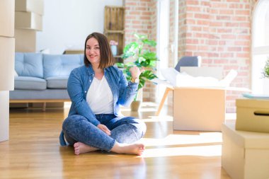 Genç ve cüsseli bir kadın karton kutuların etrafında oturmuş mutlu yüzüyle yeni bir eve taşınıyor ve başparmağıyla yan tarafı işaret ediyor..