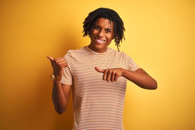 Rastalı tişörtlü Afro adam izole edilmiş sarı arka planda duruyor. Arka tarafı işaret ediyor. El ve baş parmakları havada, kendine güvenen.
