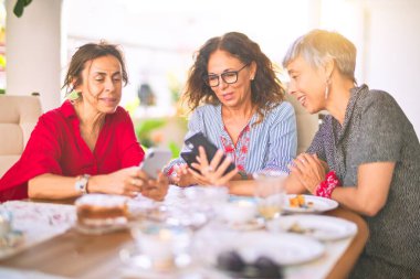 Orta yaşlı kadınlarla buluşup, öğle yemeği yiyip, kahve içiyorlar. Olgun arkadaşlar mutlu bir şekilde gülümsüyor. Güneşli bir günde evde akıllı telefon kullanıyorlar.