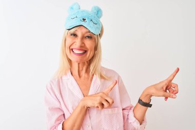 İzole edilmiş beyaz arka plan üzerinde mavi uyku maskesi ve pijama giyen orta yaşlı bir kadın gülümseyip kameraya bakıyor ve iki eliyle ve parmaklarıyla yan tarafı işaret ediyor..