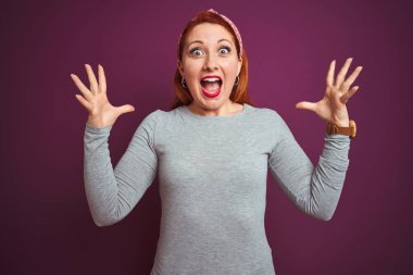 Gri tişört giyen güzel, kızıl saçlı bir kadın ve izole edilmiş mor arkaplan üzerine tepe takmış çılgın ve başarıyı kutlayan kolları açık, gözleri açık heyecanlı bir şekilde haykırış. Kazanan konsept