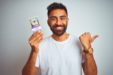 İzole edilmiş beyaz arka planda dolarlar tutan genç Hintli adam başparmağıyla gülümseyerek yüzünü gösteriyor.