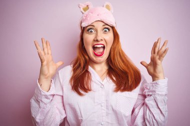 Pembe izole edilmiş arka plan üzerinde pijama giyen genç ve güzel bir kızıl kadın çılgın ve başarıyı kollarını kaldırıp heyecanla haykırarak kutluyor. Kazanan konsept
